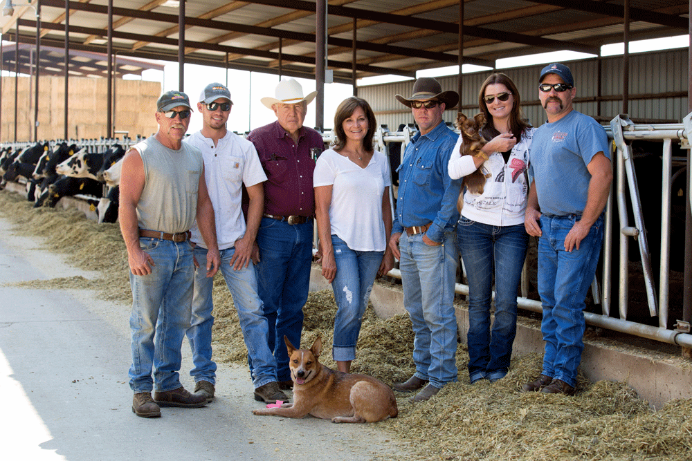 The Docheff family and their farm dogs pose for a picture while cows eat in the background.
