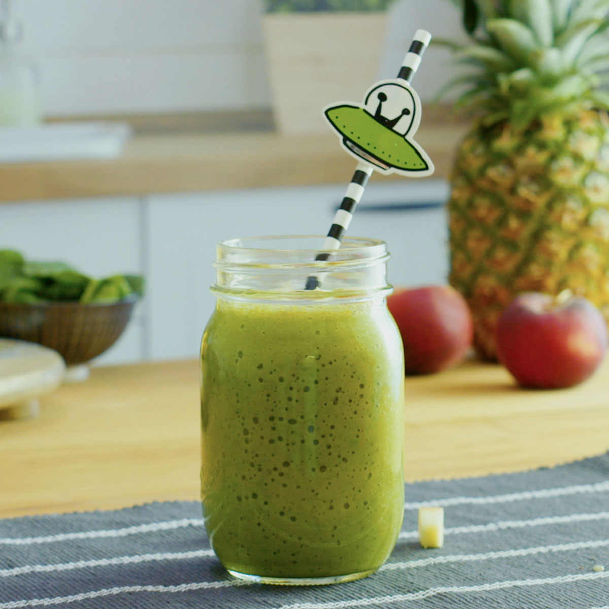 green smoothie with a fun straw on a counter with banana, pineapple in the background